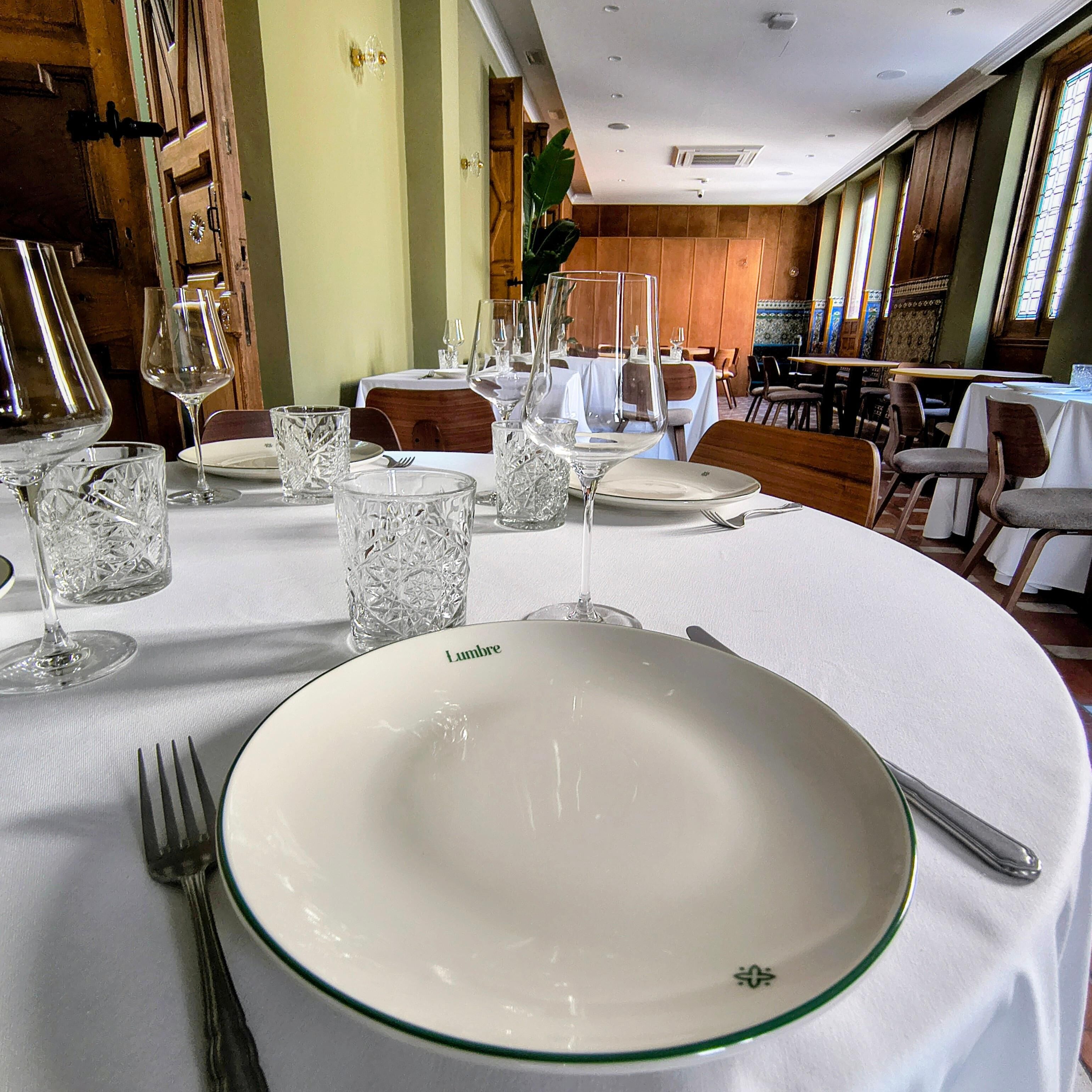 Mesa de jantar com toalha branca, prato com a etiqueta 'Lambre' e copos de vidro transparente.
