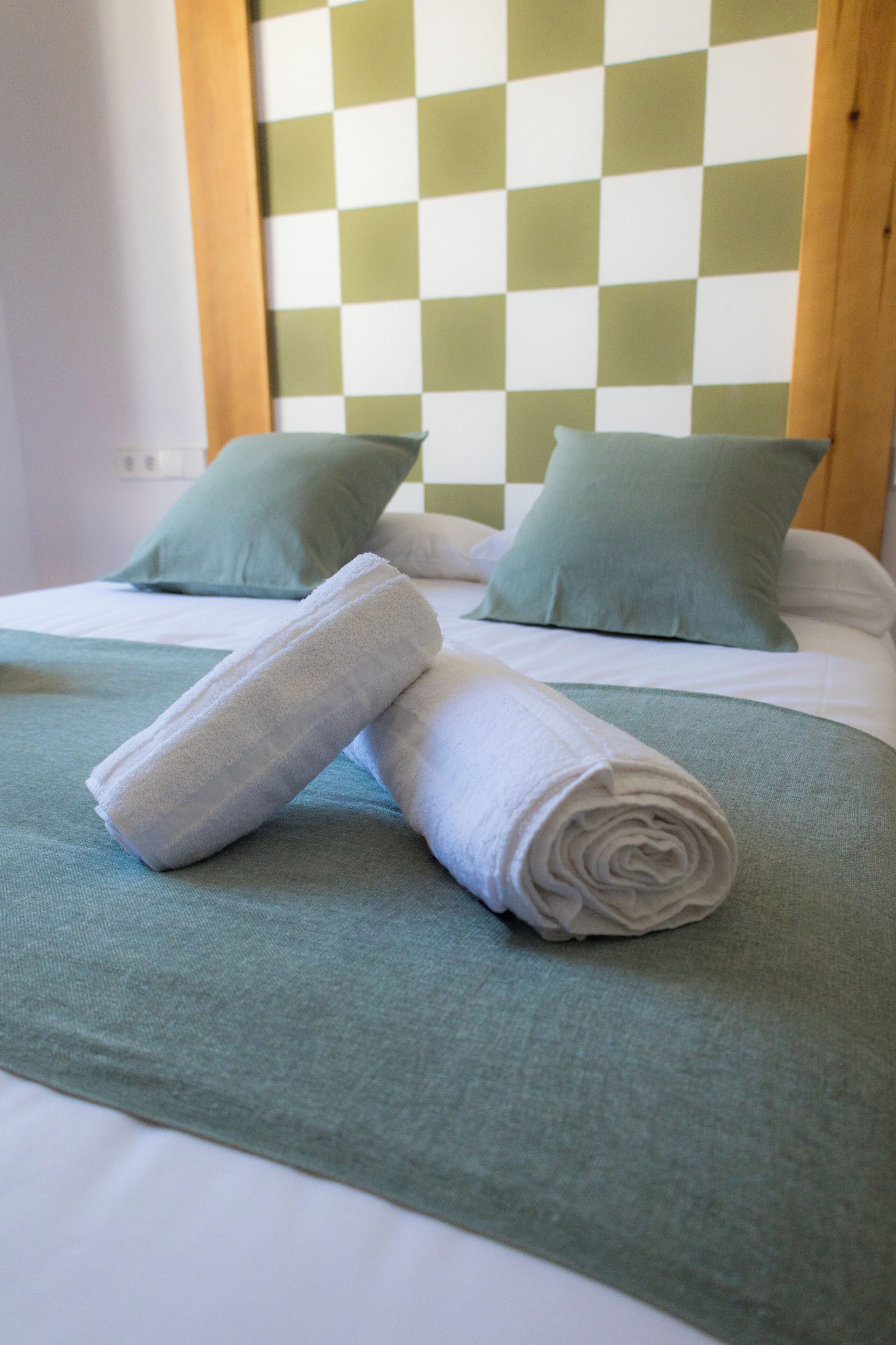 Una cama bien hecha con ropa de cama y almohadas verdes, una toalla blanca enrollada sobre la cama y un papel tapiz de cuadros al fondo.