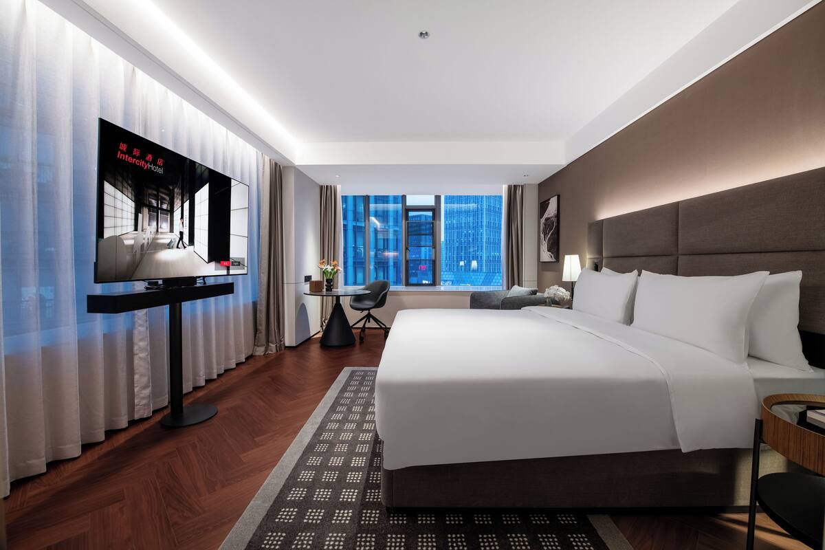 A modern hotel room with a large bed, a television, a desk, and a view of the cityscape.