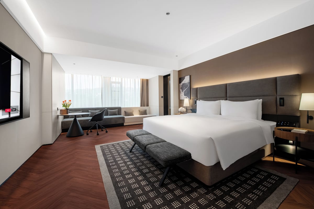 A modern hotel room with a large bed, a desk, a sofa, and a view of the city.