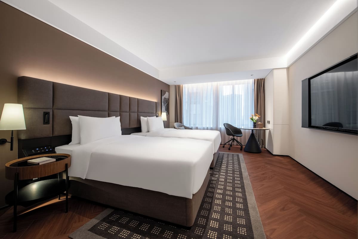 A hotel room with two beds, a desk, a chair, a TV, and a window with curtains.