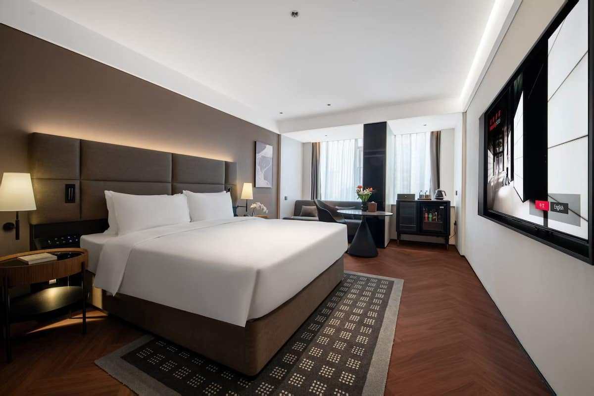 A modern hotel room with a large bed, a desk, a sofa, and a view of the city.