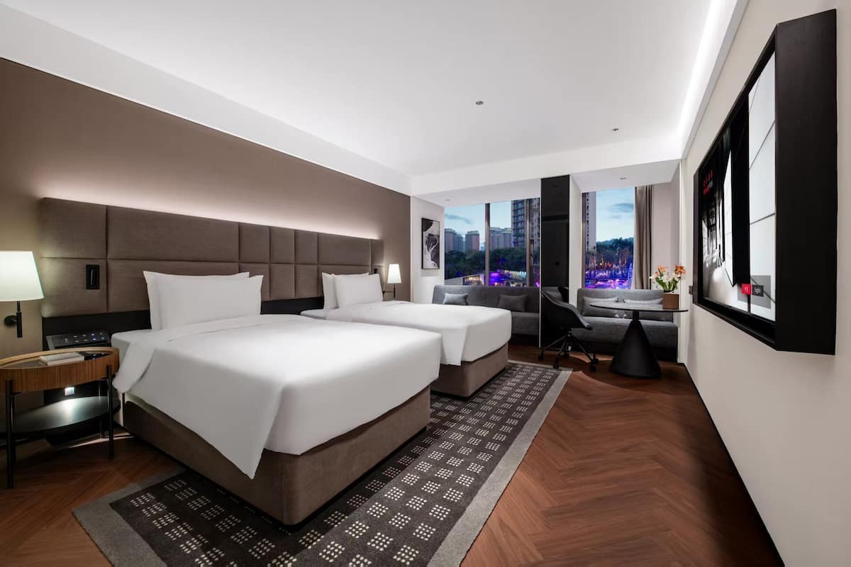 A modern hotel room with two beds, a sofa, a desk, and a view of the cityscape.