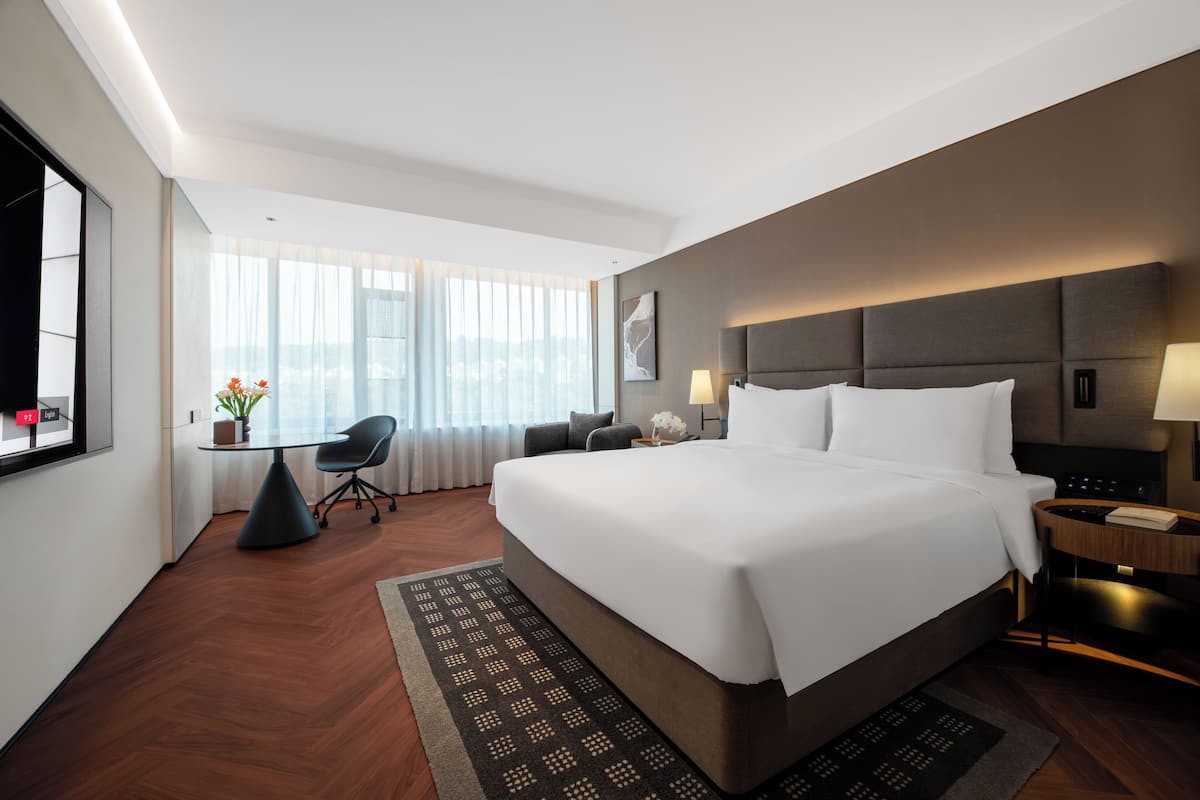 A hotel room with a large bed, a desk, a chair, a window with curtains, and a view of the city.