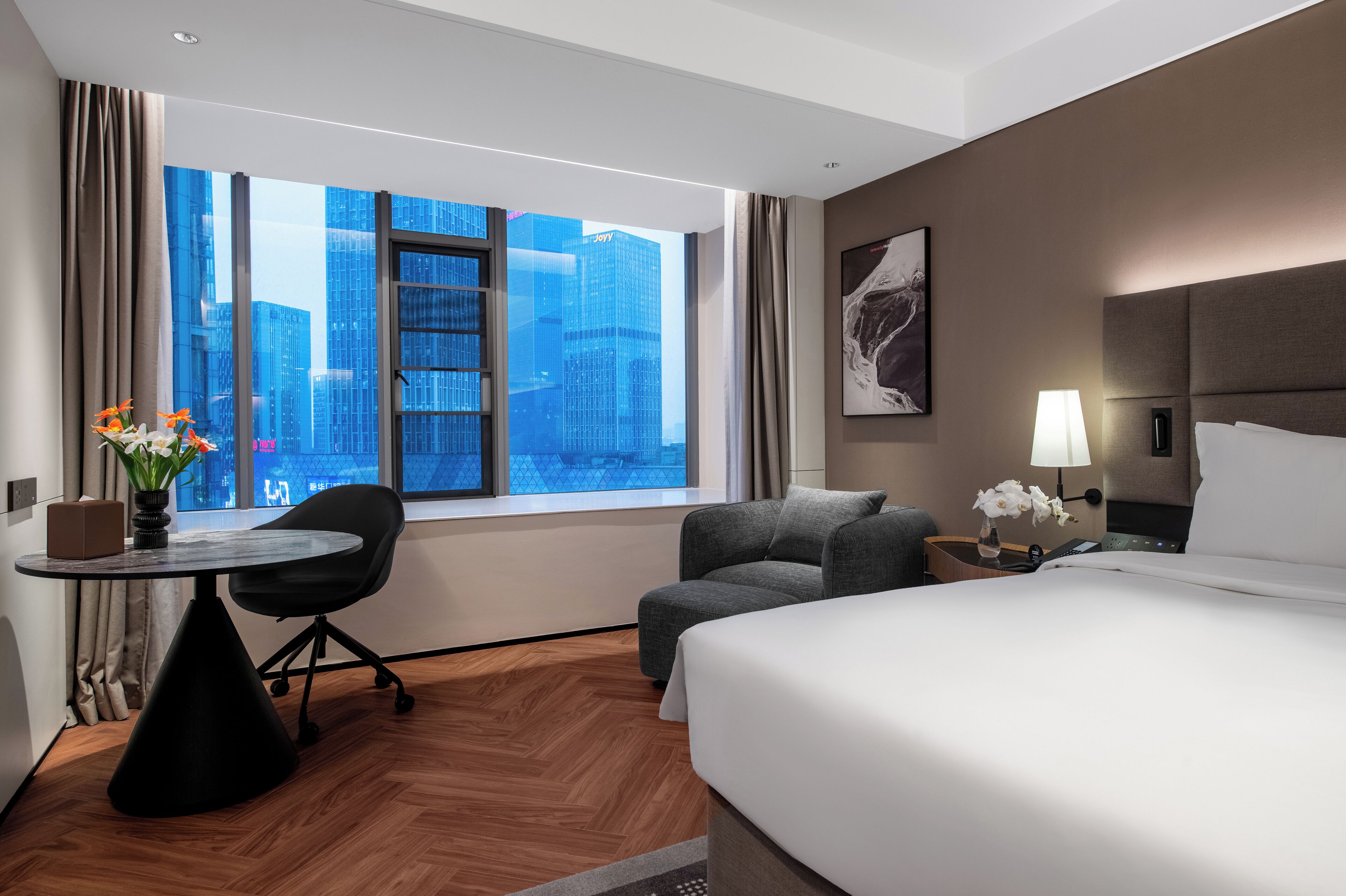A hotel room with a large window, a bed, a desk, a chair, and a nightstand with a lamp.