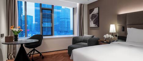 A hotel room with a large window, a bed, a desk, a chair, and a nightstand with a lamp.
