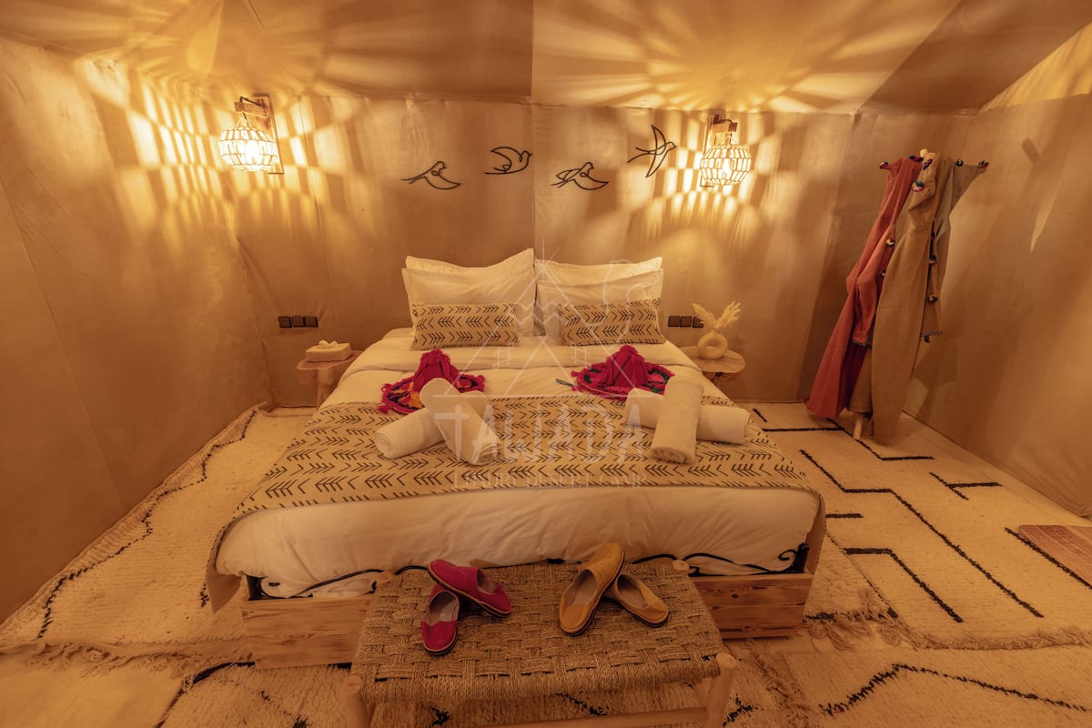 Un dormitorio con cama, almohadas, mesita de noche, un cofre de madera y una lámpara fijada a la pared.
