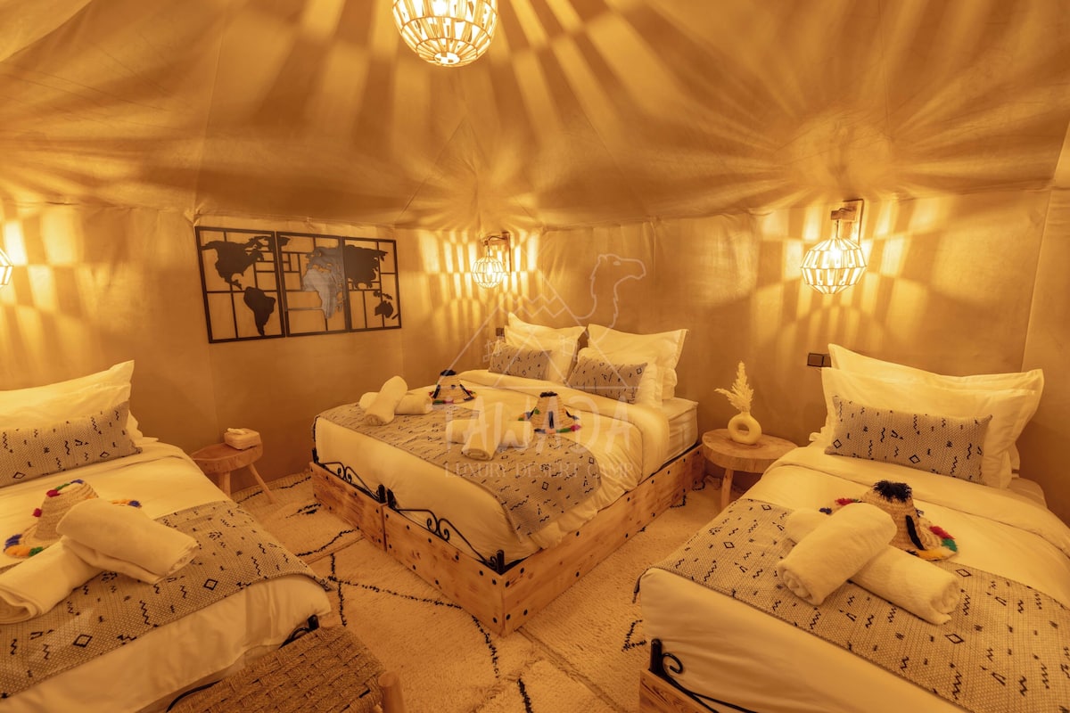 Habitación de hotel con una cama grande, dos camas más pequeñas, una mesita de noche, una lámpara y un cuadro en la pared.
