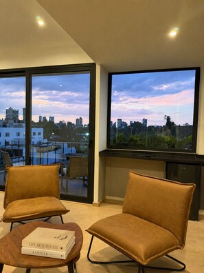 Ein modernes Wohnzimmer mit Blick auf die Stadt, zwei braune Ledersessel, ein Couchtisch mit Büchern und ein großer Flachbildfernseher.