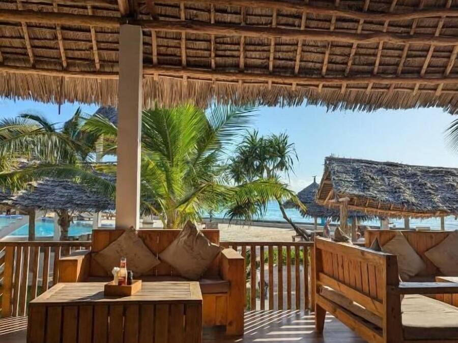Una terraza de madera con asientos, techo de paja y vistas a una piscina y palmeras.