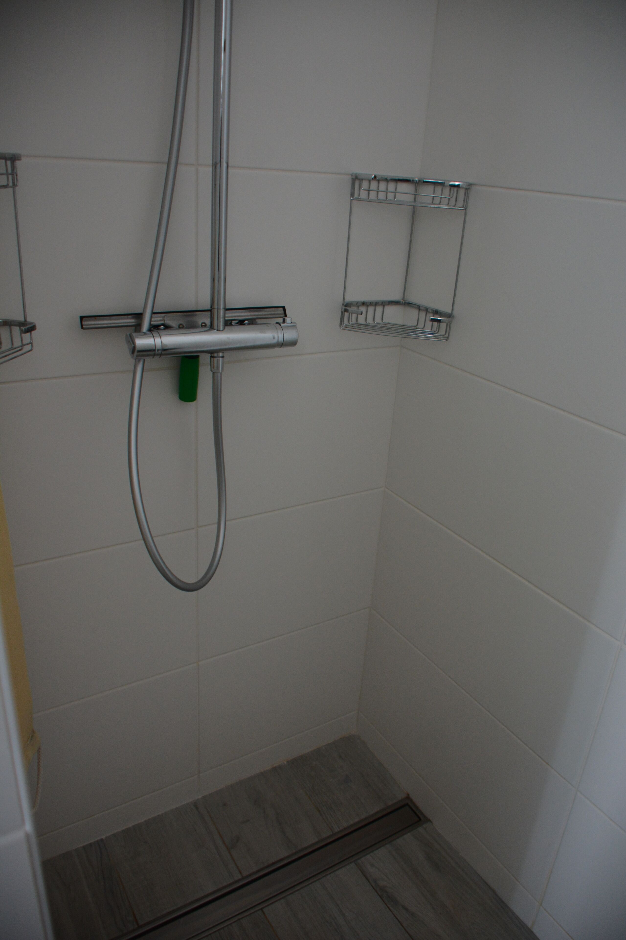 Une douche équipée d’un pommeau de douche à main et d’un robinet mural, une petite étagère pour les articles de toilette et un plancher en bois.