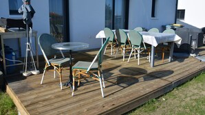 Eine Holzterrasse mit Gartenmöbeln, darunter ein runder Glastisch, Stühle und ein Grill.