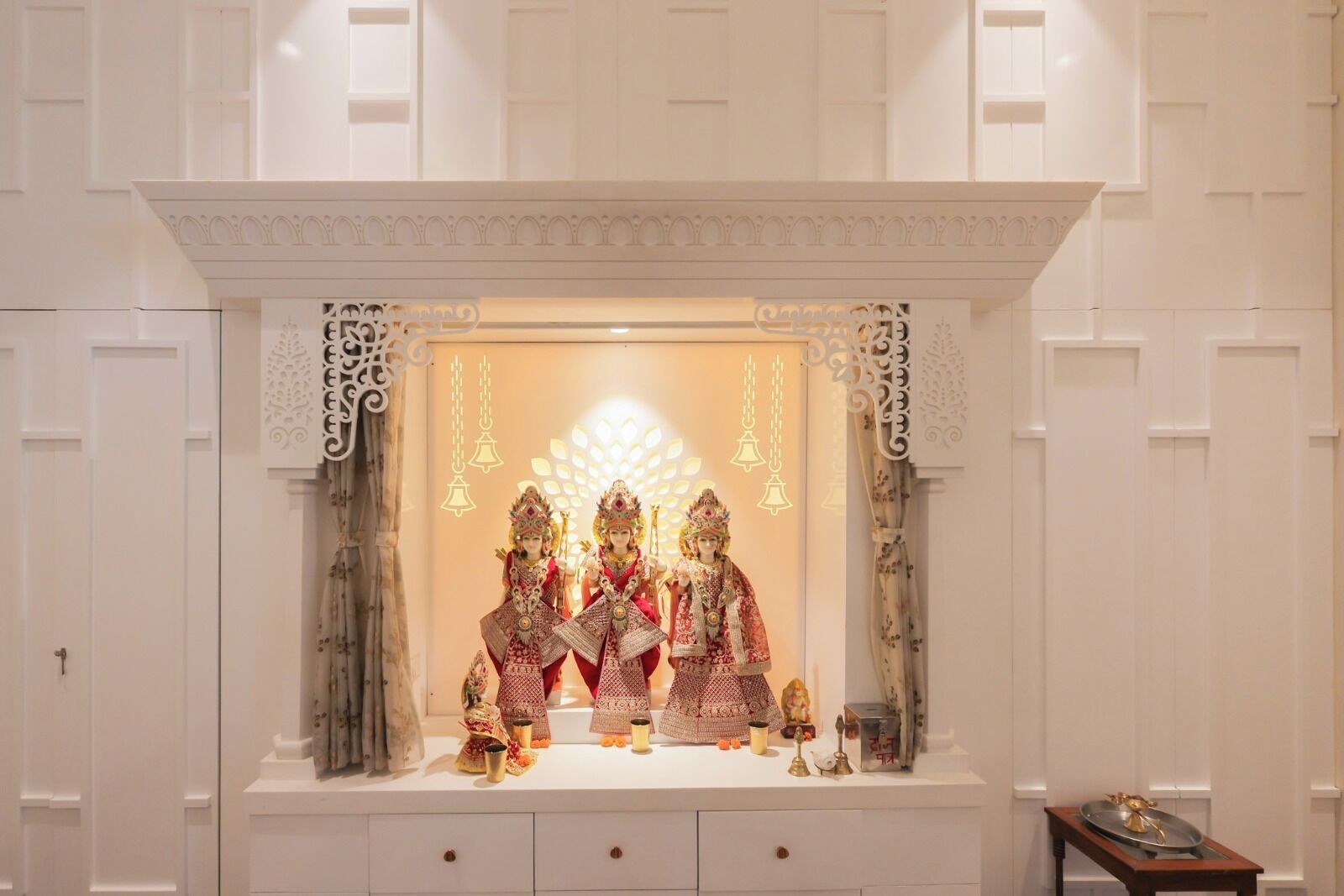 Un sanctuaire orné de trois statues, un mur blanc avec des panneaux décoratifs et une petite table sur laquelle sont posés divers objets.