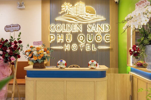 A reception desk with a neon sign for Golden Sand Phu Quoc Hotel, a wicker basket with flowers, and a wooden counter with a plant.