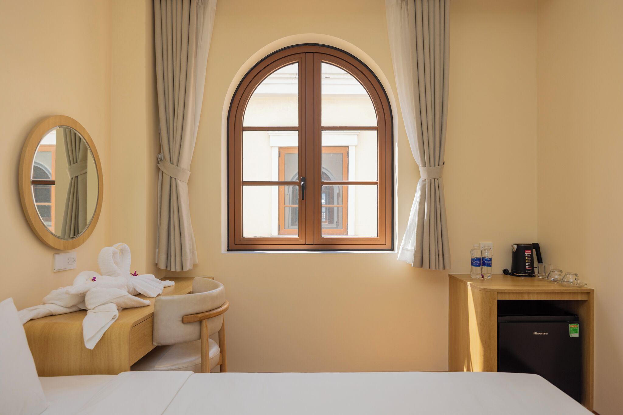 A hotel room with a bed, a wooden desk with a sink, a mirror, a window with curtains, and a minibar.