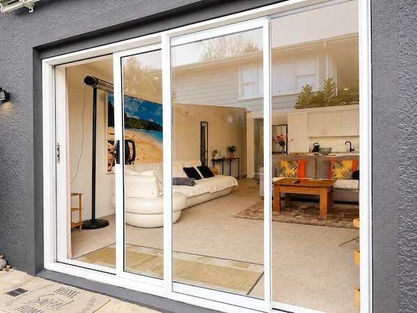 A modern home interior with a sliding glass door leading to a living room, a sofa, a coffee table, and a kitchen area visible through the glass.