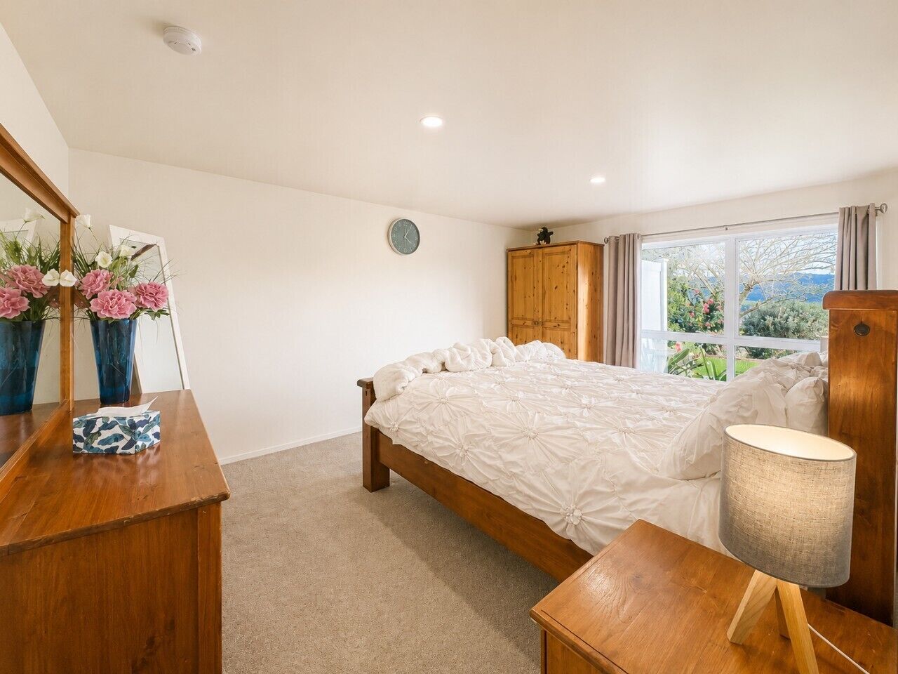 A bedroom with a bed, a wooden dresser, a bedside table, a lamp, a mirror, and a window with curtains.
