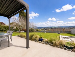 A patio with a table and chairs, a covered area, a grassy yard, and a view of hills and a fence. - Stylish 1BR Retreat | Patio | Walk to Shops | Parking (Whangārei)