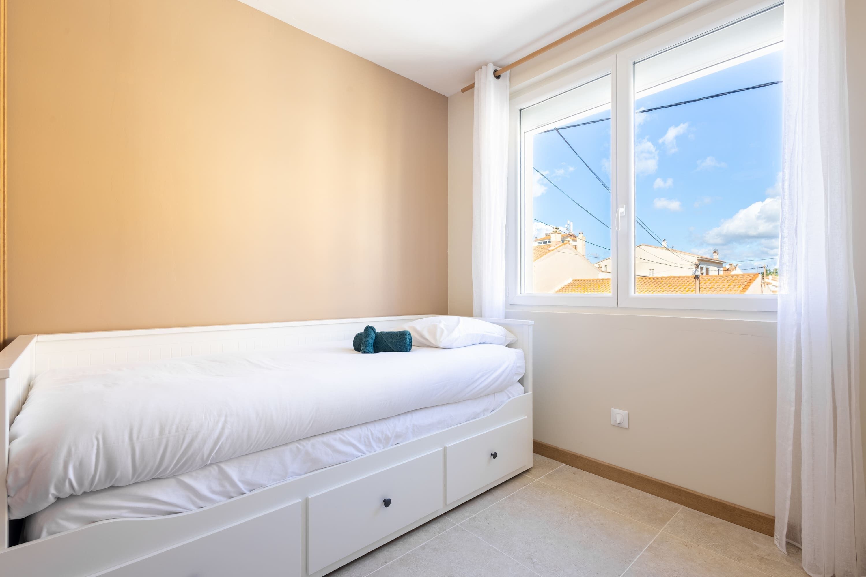 Une chambre avec un lit simple, une fenêtre avec des rideaux et une vue sur les bâtiments environnants.