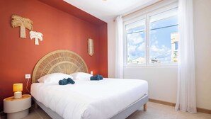 Ein ordentlich bezogenes Bett mit weißen Leinen, ein runder gewebter Nachttisch mit Lampe und ein Fenster mit Blick auf Gebäude.