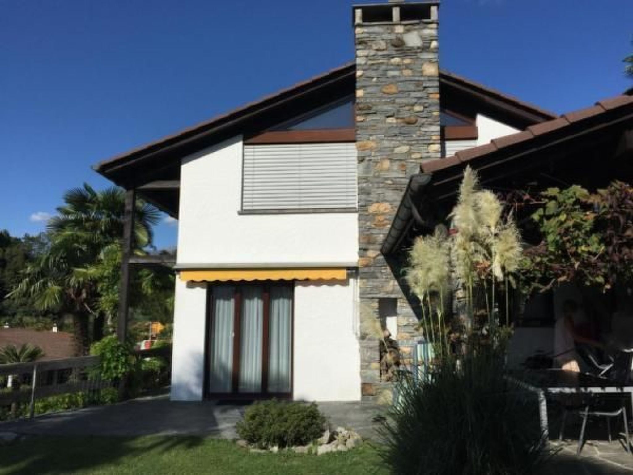 Uma casa moderna de dois andares com chaminé de pedra, fachada branca e um pequeno jardim frontal com plantas.