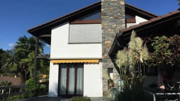 Uma casa moderna de dois andares com chaminé de pedra, fachada branca e um pequeno jardim frontal com plantas.