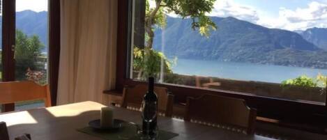 A dining table with chairs, a candle, and a bottle, set against a window with a view of mountains and a lake.