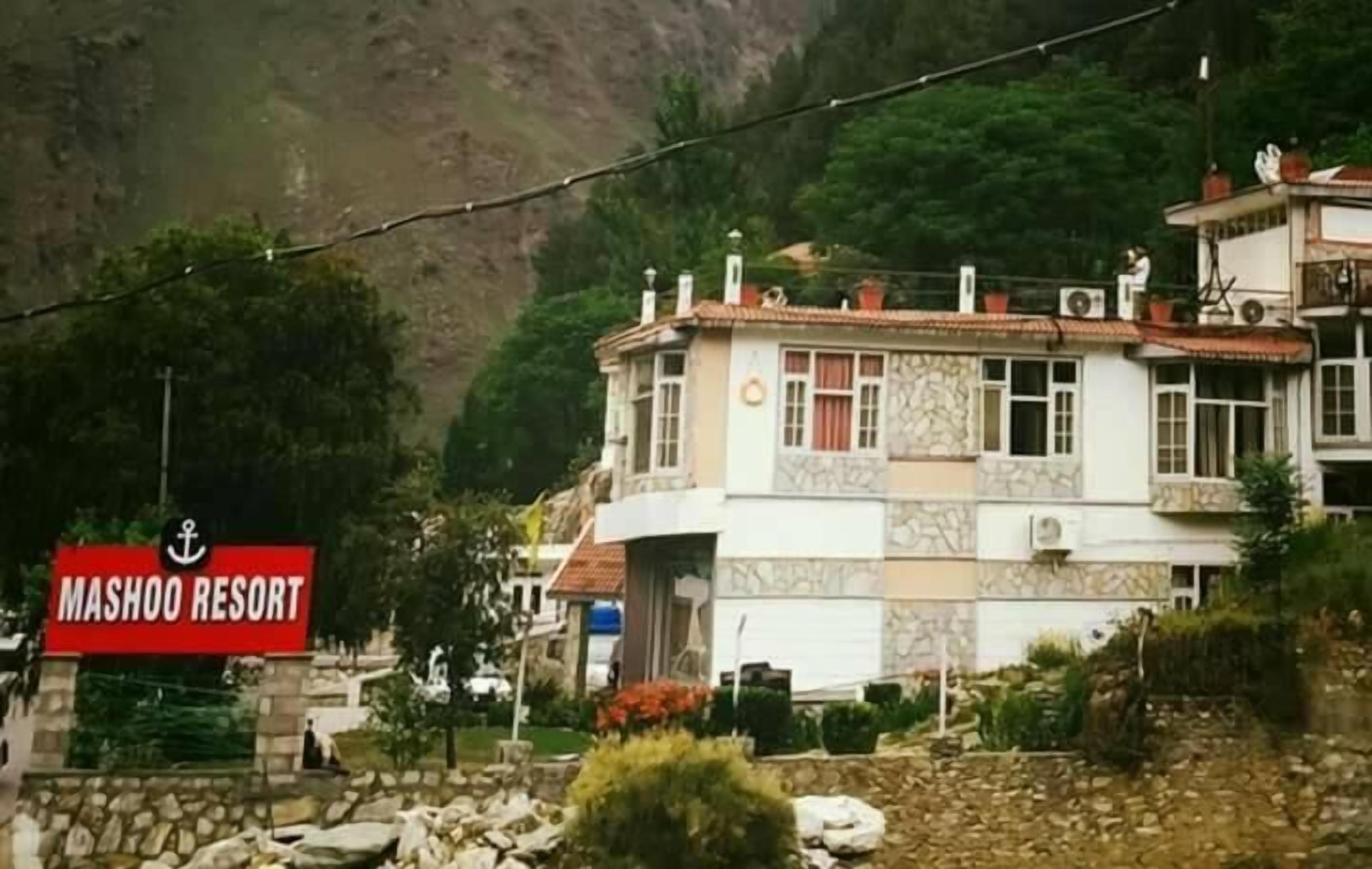 Un bâtiment sur lequel on peut lire l’inscription « MASHOO RESORT » sur une enseigne, entouré de verdure et avec une chaîne de montagnes en arrière-plan.