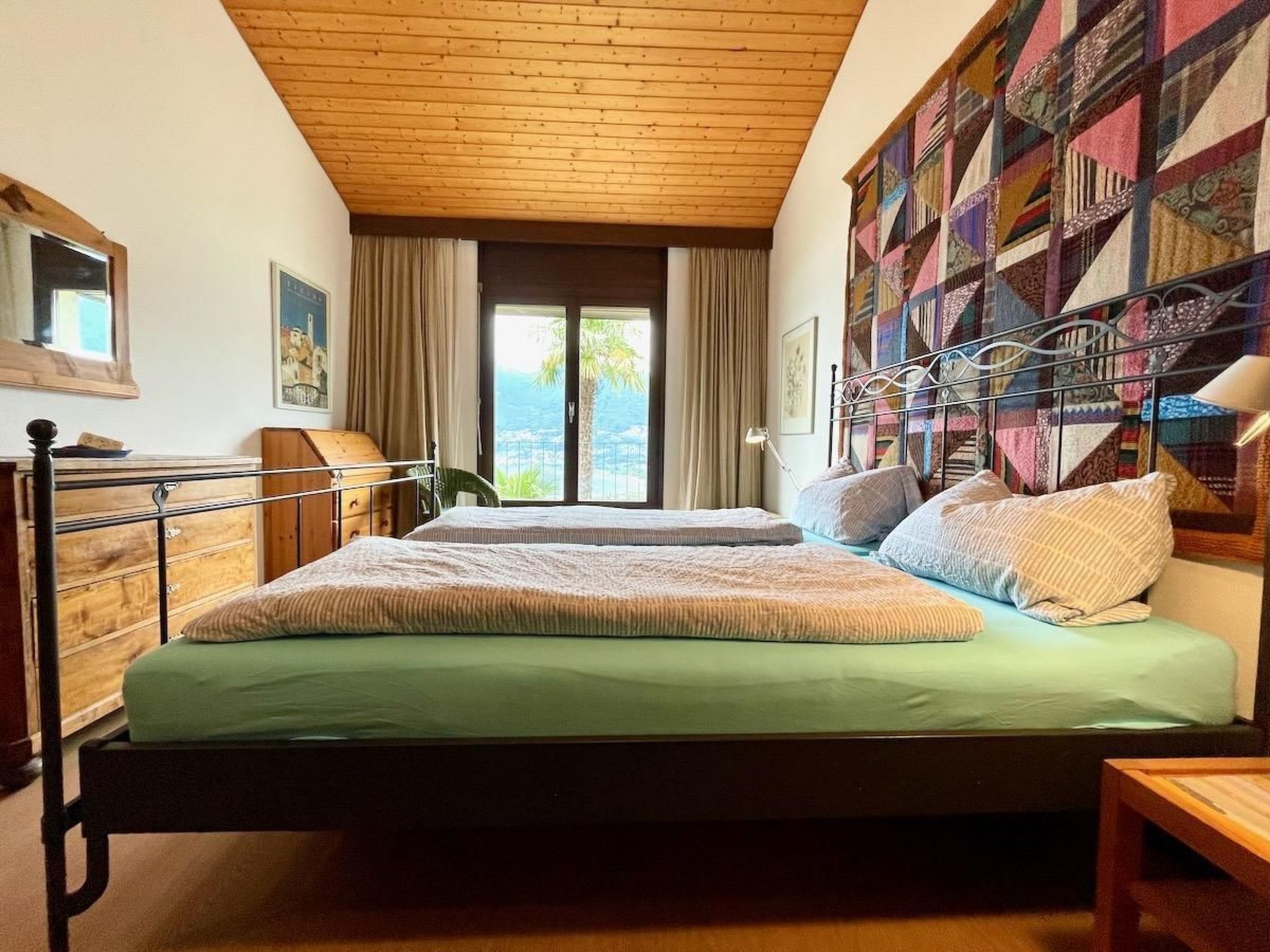 A bedroom with a bed, a desk, a chair, a window with curtains, and a view of the outdoors.