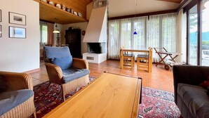 A cozy living room with a fireplace, wooden furniture, and a large window with curtains. - Casa Poggio Verde by Interhome (Magadino)