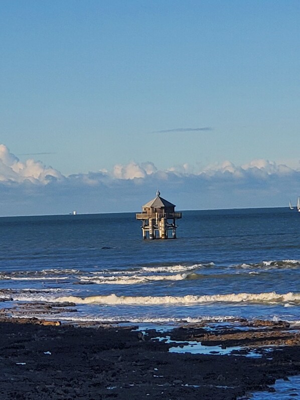 Eine Küstenlandschaft mit einer kleinen Holzkonstruktion auf Stelzen im Wasser, Wellen schlagen an eine felsige Küste.