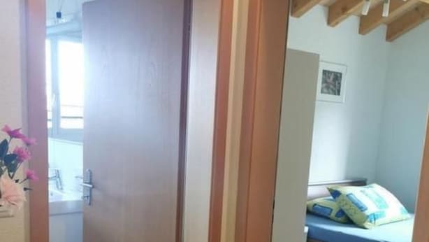Ein Zimmer mit einem Bett, einem Holztisch und einer Tür zu einem weiteren Raum.