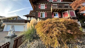 Une maison en bois à deux étages, avec des volets rouges, une statue de lion en pierre et un jardin où poussent différentes espèces de plantes.