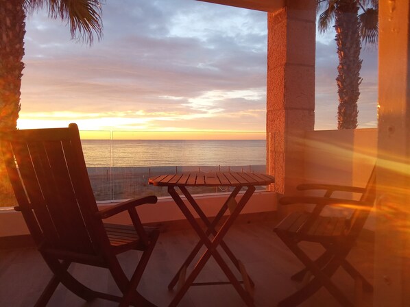 Un balcon avec une table en bois et deux chaises, d’où l’on peut admirer le coucher de soleil sur l’océan.