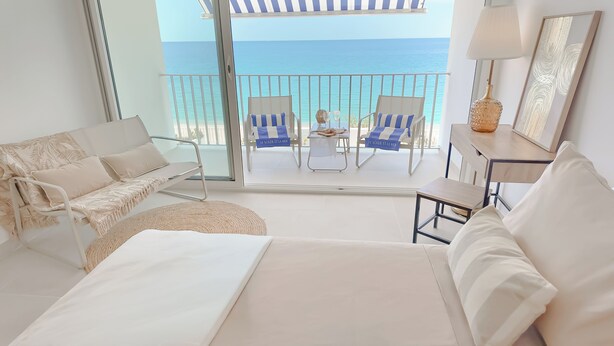 A hotel room with a bed, a sofa, a small table, and a chair, all with white bedding. There is a balcony with a view of the sea. - Luxury Beachfront Studio - Panoramic sea view - Direct beach access - Corsica (Olmeto)