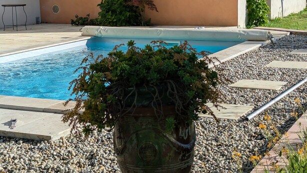 A swimming pool with a stone border, a large decorative urn, and a building with a peach-colored wall and a window. - Gîte avec piscine privée situé à Uzès (Uzès)