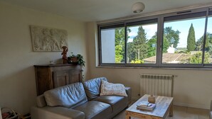 A living room with a leather sofa, a wooden coffee table, a radiator, and a window with a view of trees and buildings. - Gîte avec piscine privée situé à Uzès (Uzès)