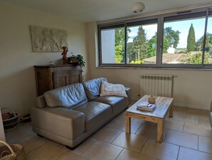 A living room with a leather sofa, a wooden coffee table, a radiator, and a window with a view of trees and buildings. - Gîte avec piscine privée situé à Uzès (Uzès)