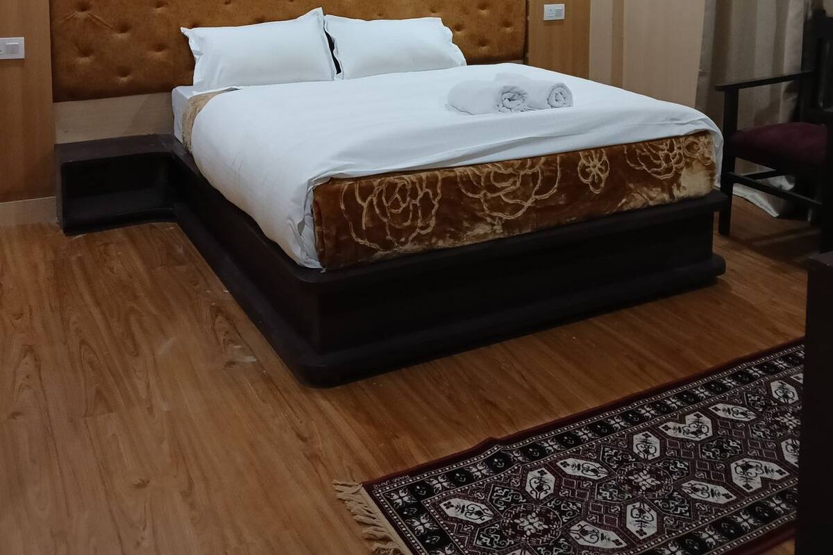 A bedroom with a large bed, a headboard, a wooden floor, a patterned rug, and a ceiling light.