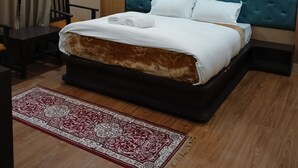 A neatly made bed with a blue headboard, a wooden nightstand, a chair, and a patterned rug on a wooden floor. - Hotel Shree Jee Inn (Badrinath)