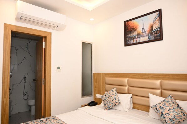 Una habitación de hotel con cama, cabecera de madera, cuadro de un paisaje urbano, baño con pared de mármol y unidad de aire acondicionado.