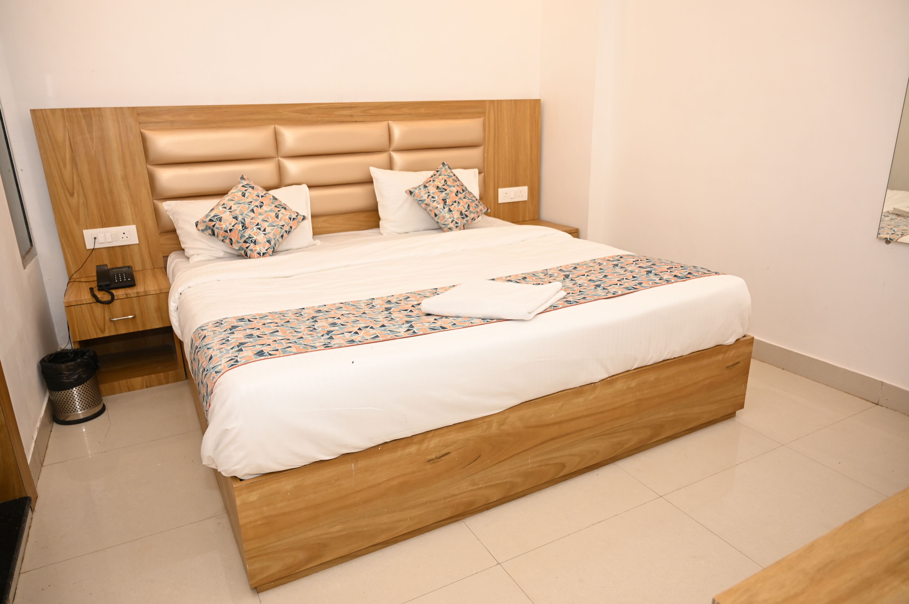 A neatly made bed with a wooden headboard, a bedside table with a phone, and a trash can.
