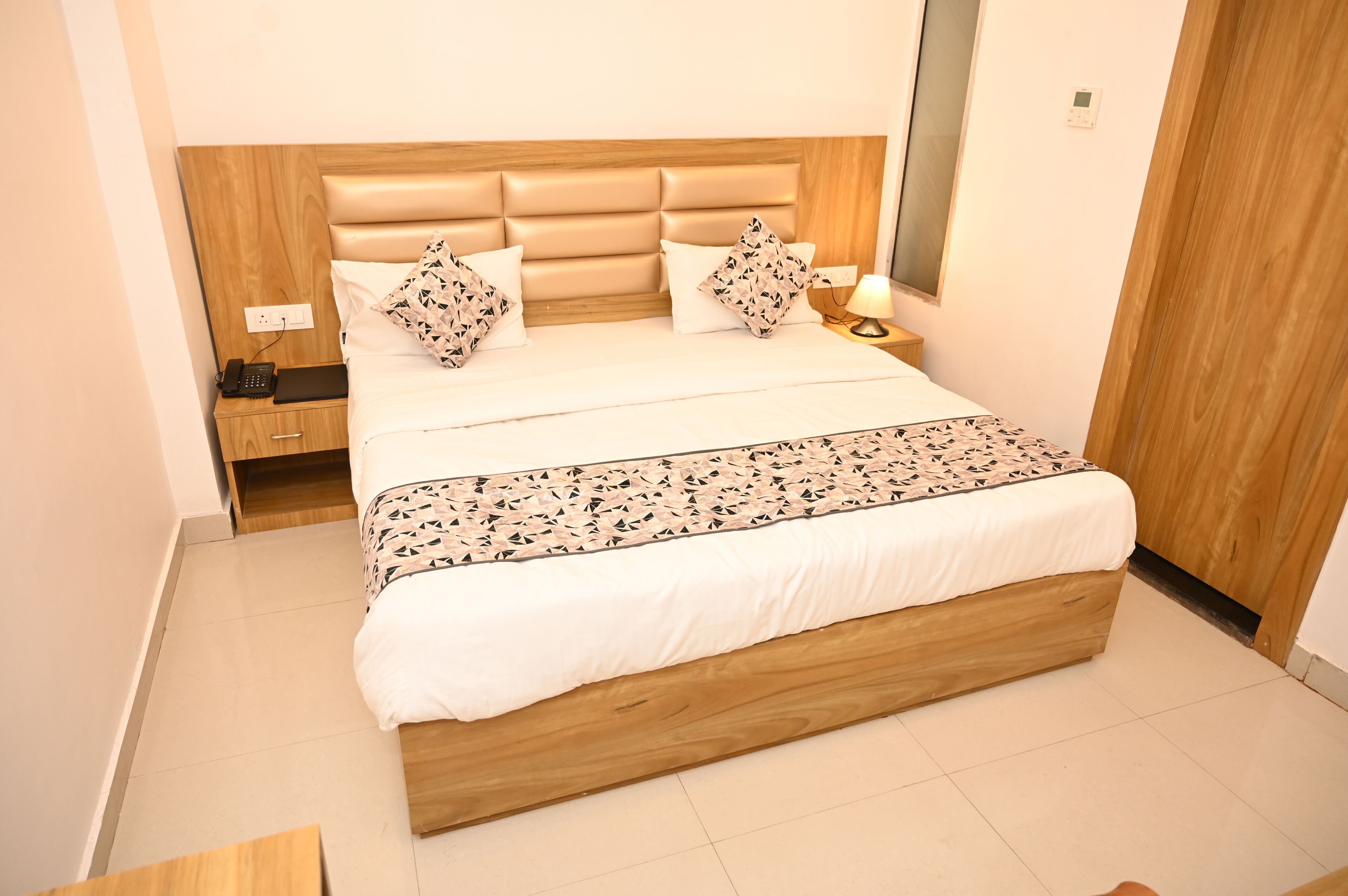 Una cama bien tendida con cabecera de madera, una mesita de noche con lámpara y un enchufe.