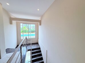 Una scala con ringhiera in metallo, una finestra vista su verde e una recinzione, e un soffitto bianco con faretti.