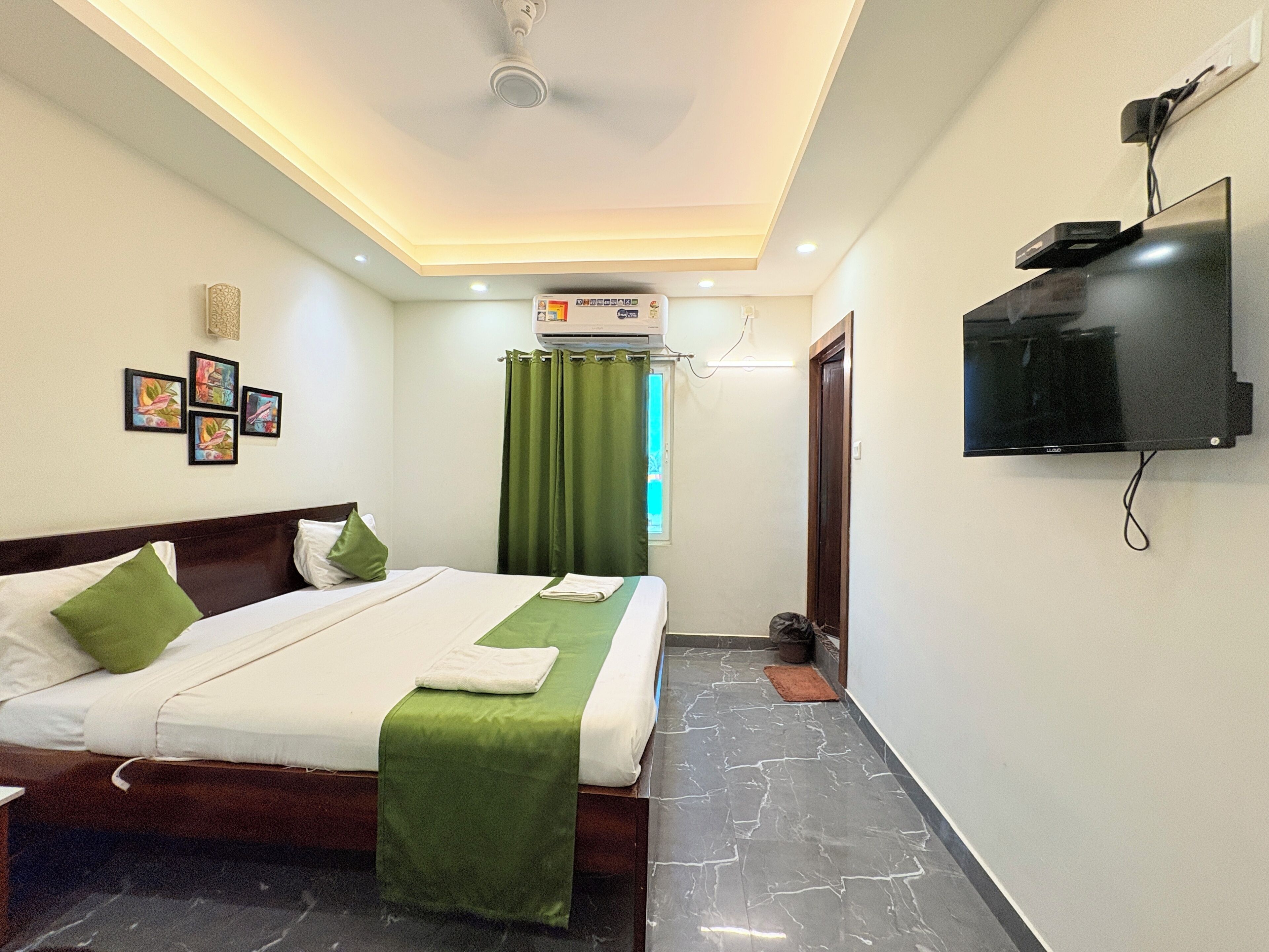 A hotel room with a bed, a flat-screen TV mounted on the wall, a ceiling fan, and a window with curtains.