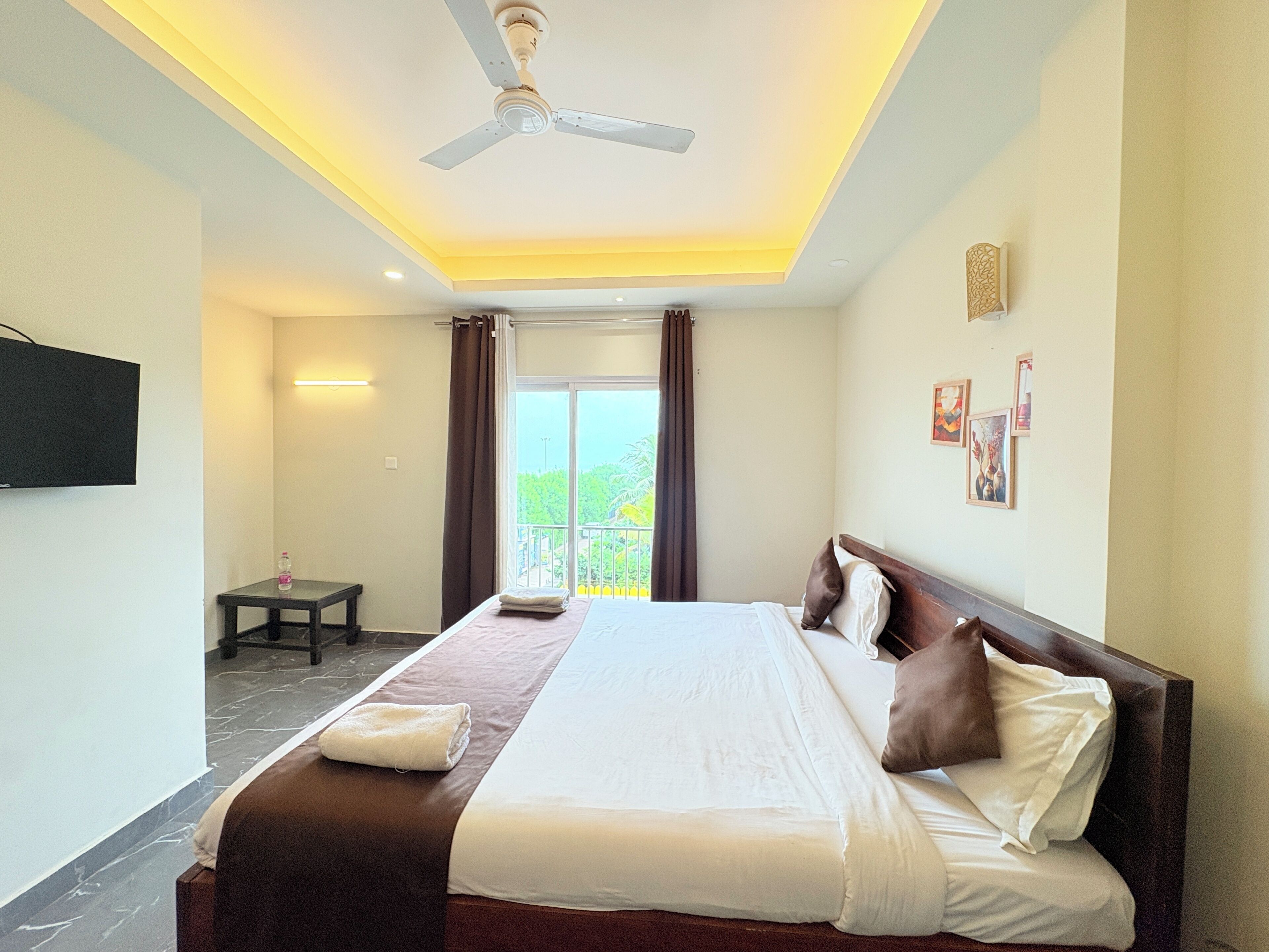 A hotel room with a bed, a ceiling fan, a TV, a small table, and a view of greenery through the window.