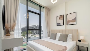 Ein modernes Hotelzimmer mit großem Fenster, einem Bett mit vielen Kissen, einem Nachttisch mit Lampe und zwei gerahmten Kunstwerken an der Wand.
