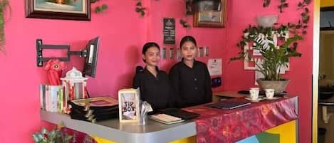 Un mostrador de recepción de colores brillantes con un reloj digital, dos empleados y un diseño floral en la pared.