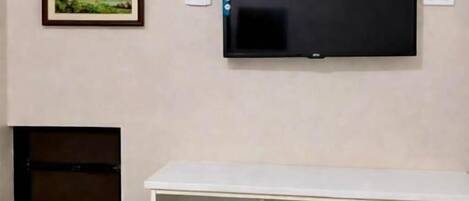 Una habitación con una televisión de pantalla plana montada en la pared, un escritorio blanco, dos sillas de madera y un refrigerador pequeño.