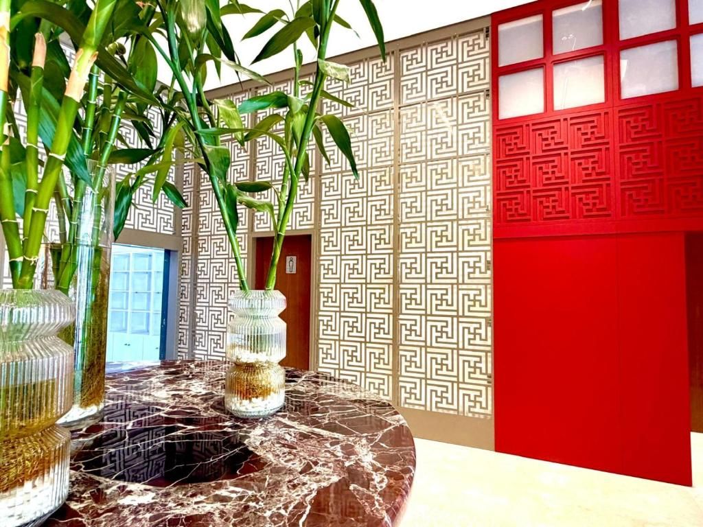 A room with a marble table, two glass vases with plants, a patterned wall, and a red door.